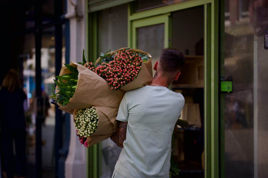 A person with short dark hair, wearing a light grey T-shirt, is seen from behind carrying a large bouquet of fresh flowers in their right arm. The bouquet includes clusters of pink berries, white berr