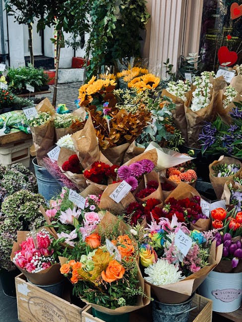 A vibrant display of fresh flower bouquets arranged outdoors on wooden crates and metal buckets, featuring a variety of blooms including bright yellow sunflowers, pink lilies, orange roses, purple iri