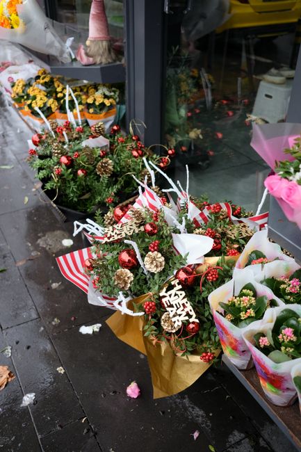 A festive floral arrangement featuring a bouquet of fresh evergreen branches, red berries, pinecones, and shiny red baubles, wrapped in brown paper and decorated with white ribbons. The bouquet is pla