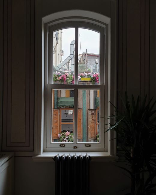 A view through a large, arched window with white framing, showcasing an outdoor balcony decorated with vibrant pink and purple petunias in flower boxes. The windowpane reveals a glimpse of neighboring