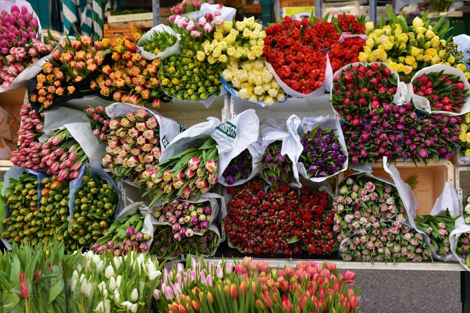A vibrant display of freshly cut flower bouquets arranged on shelves at a flower market, showcasing a variety of tulips, roses, and other blooms in shades of pink, red, yellow, purple, and white. The