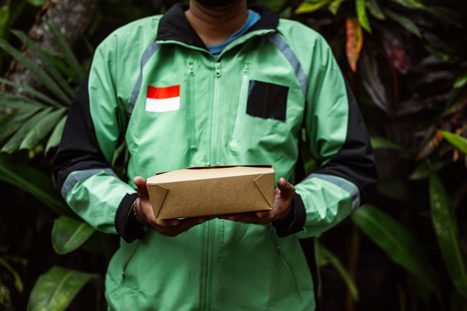 A delivery person from [COMPANY_NAME] wearing a bright green jacket with black accents and a red and white flag patch on the chest, is holding a rectangular cardboard parcel. The background features l