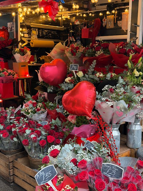 A vibrant display of floral arrangements and bouquets at a flower shop, featuring red and white roses, tulips, and mixed greenery. The flowers are wrapped in clear and pink cellophane, with some tied