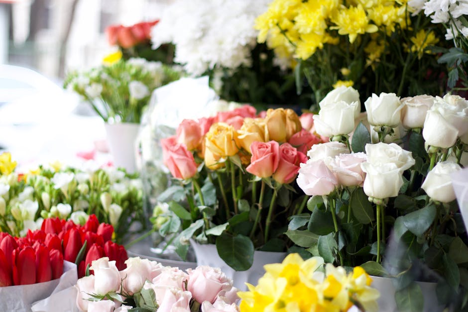 A vibrant display of fresh flowers arranged in bouquets at a flower shop, including white, pink, yellow, and red roses and tulips. The flowers are arranged in wrapping paper or placed in vases and pos