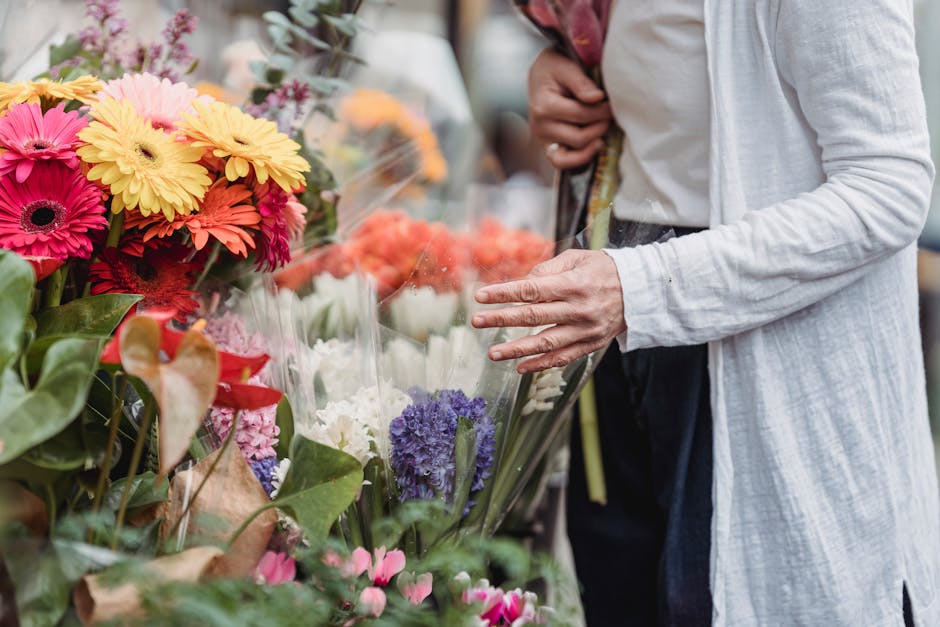 A person in a white long-sleeve top selecting fresh flowers at a floral market stall. The bouquet includes vibrant pink, yellow, orange, and purple blooms, such as daisies, hydrangeas, and anthuriums,