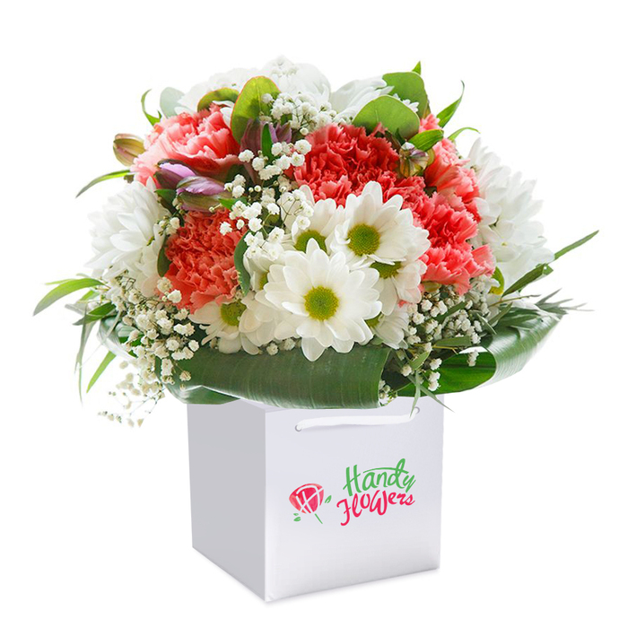 Presented upright in a crisp, contemporary white gift bag, this expertly crafted bouquet features an abundant blend of soft coral, peach, and radiant red carnations-all marked by their exquisitely ruffled petals that add richness and depth. Scattered among the carnations are pristine white daisies, each with a lively lime-green center reminiscent of the vibrant flower stalls along Kentish Town High Street. Delicate sprays of white baby's breath interlace delicately between the larger blooms, creating graceful interludes and an airy, open feel that contrasts beautifully with the deep hues of the main flowers. Along the lower edge, lush, broad-leaved greenery wraps the arrangement in a natural collar, their glossy depths adding structure and stability, much as Hampstead Heath borders the neighbourhood's busy streets. Not to be overlooked, a few unopened purple-tinged buds-perhaps promising alstroemeria-nestle on the left side, hinting at new beginnings and creating additional visual interest. The dual ribbon handles of the white bag arch gently at the top for comfortable carrying, the subtle branding on the front blending seamlessly into the refined composition. Shot against an immaculate white background, the lighting highlights every rich hue and soft petal edge, underlining the bouquet's readiness for same-day delivery. This arrangement, assembled with expertise and a keen understanding of Kentish Town's vibrant community spirit, is perfect for special events like birthdays or anniversaries-or any moment deserving a thoughtful gesture rooted in local care and craftsmanship.