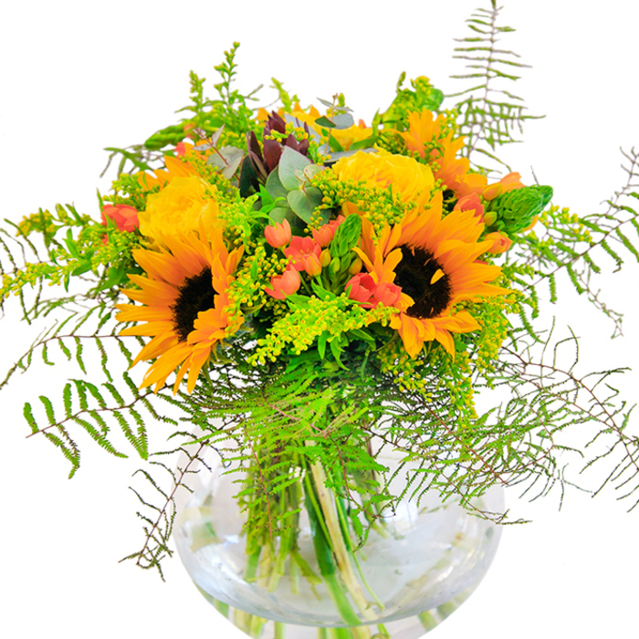 Upon first glance, this arrangement is a vivid celebration of Kentish Town's lively spirit, bursting with the radiant yellows of sunflowers and garden roses that recall the sun-drenched corners of Highgate Road. The sunflower heads form the beating heart of the bouquet, their dark centers and golden fringes drawing the eye in. Around them, tightly packed yellow rose rosettes unwind, each layer of petals exquisitely arranged, capturing both classic elegance and cozy familiarity. Clusters of deep red Hypericum berries are artfully woven throughout, gleaming amidst lush, glossy foliage and providing a peppering of rich contrast. The composition is softened by arching, feathery fern fronds and sprays of delicate green filler-possibly goldenrod-peppered with tiny yellow florets that twinkle in the natural light. Sprigs of eucalyptus tuck in at intervals, cool and aromatic, adding complexity to the green palette. The bouquet is cradled within a crystal-clear spherical vase, its contents gleaming and reinforcing a sense of freshness, like a breath of early morning air drifting in from Dartmouth Park. This detailed, sensory arrangement delights as a centerpiece or cheerful gift, offering a harmonious blend of color, scent, and texture that encapsulates the easygoing charm of everyday life in Kentish Town.